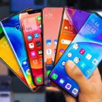 A hand holding six different phones in front of a wall.