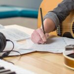 A person writing on paper with a guitar and headphones