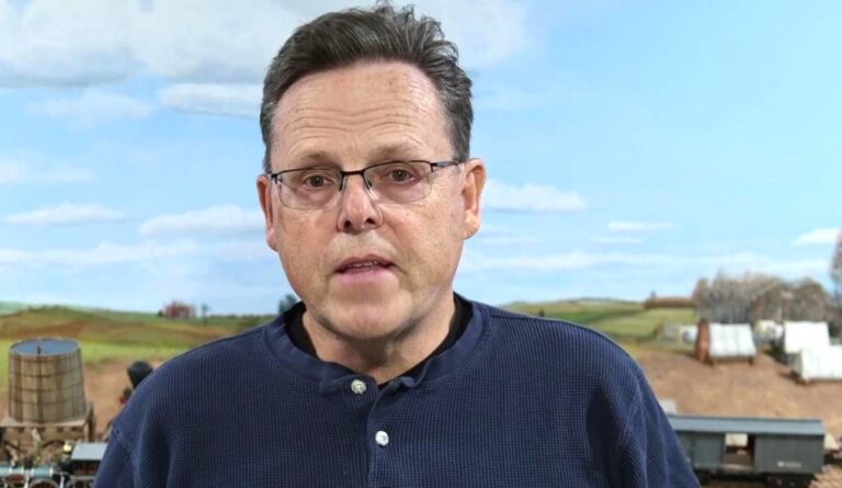 A man with glasses is standing in front of a field.